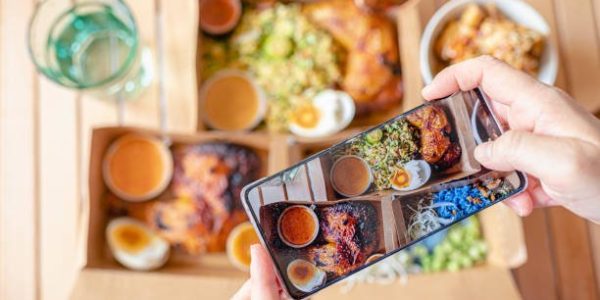 Personal perspective human hand taking photo using smart phone on table top view malaysian food nasi kerabu, nasi ulam and ayam percik in recycled paper container with sauce