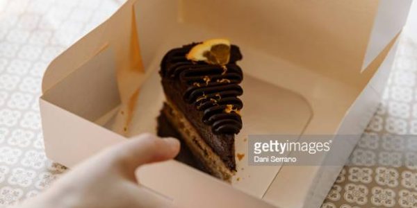 From above crop anonymous person with delicious vegan piece of chocolate cake in paper container from takeaway food delivery order at home. Sweet chocolate and orange slice of vegan cake in recyclable box.