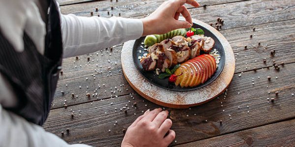 Chief decorated  baked veal on wooden table. Over the shoulder view of food decoration.  Chief decorated  dietary meat with fresh tropical fruits.